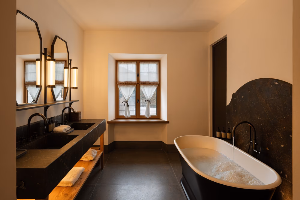 Suite bathroom at Castel Badia with a freestanding soaking tub, dark stone double vanity, and a window with mountain views