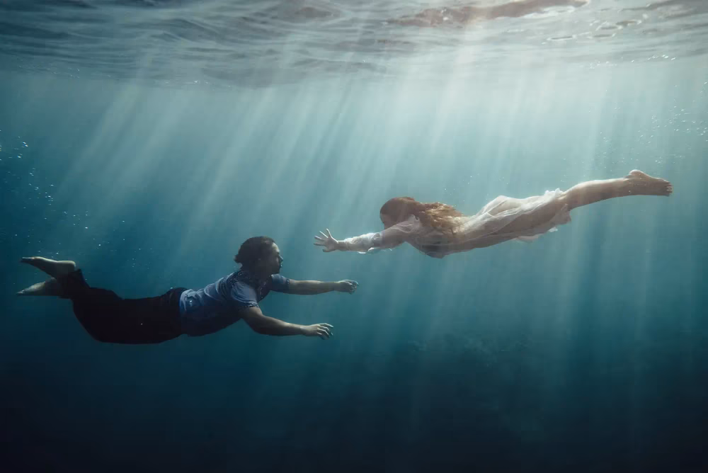 man and woman underwater swimming toward each other with sunlight coming through