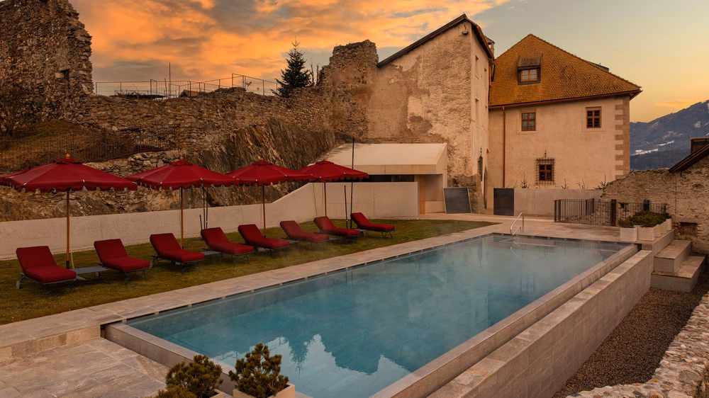 Outdoor pool with red loungers and umbrellas beside castle ruins at sunset at a Dolomites castle hotel