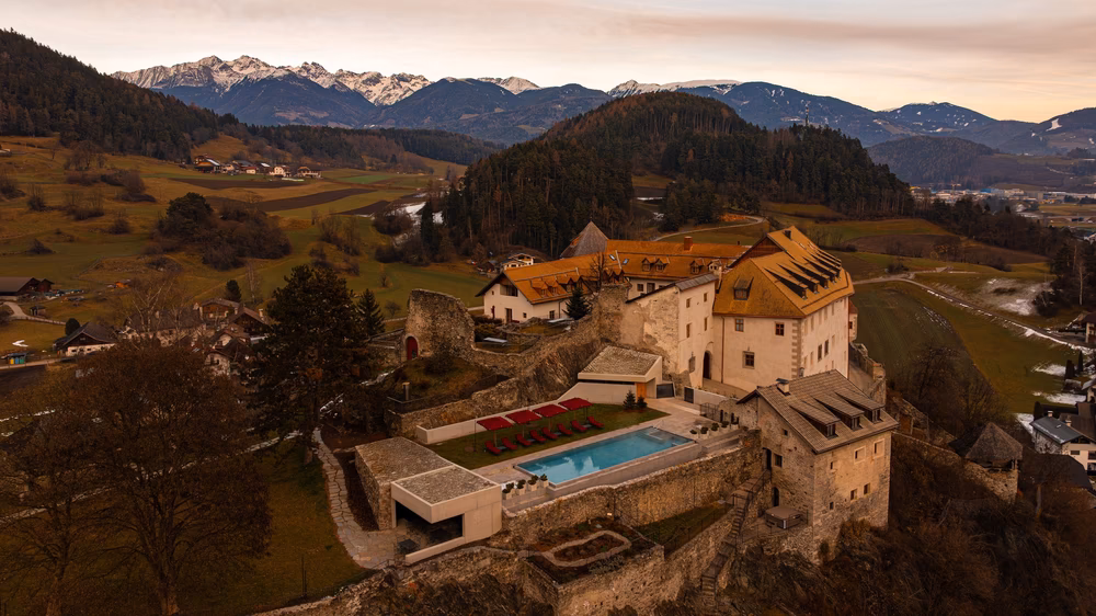 Aerial view of Castel Badia, a hilltop castle hotel and wedding venue in the Dolomites, with an outdoor pool and snow-capped peaks behind