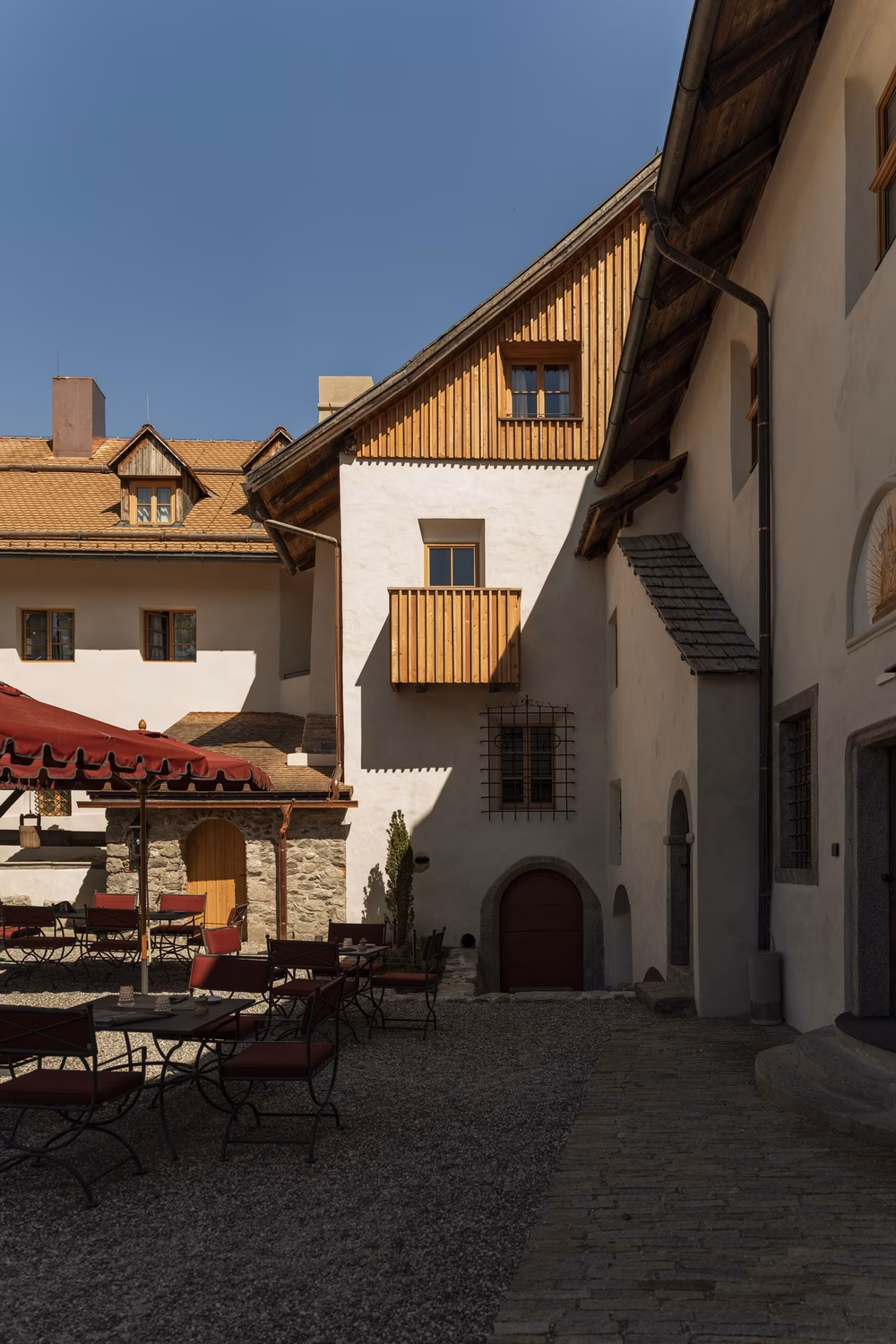 Castle courtyard at Castel Badia with white stone walls, a wooden balcony, and terrace tables under red umbrellas on a sunny spring day